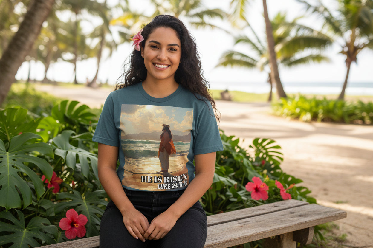 Woman wearing a t-shirt with a graphic design of a Pacific Island Jesus  on a beach, sitting on a wooden bench with tropical plants and flowers around.