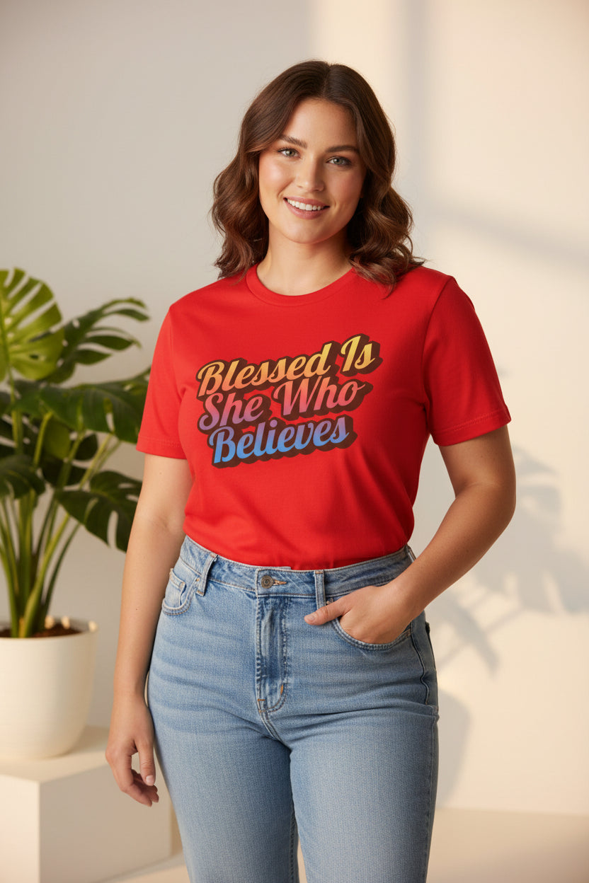 Woman wearing a red t-shirt with 'Blessed is she who believes' text, standing indoors.